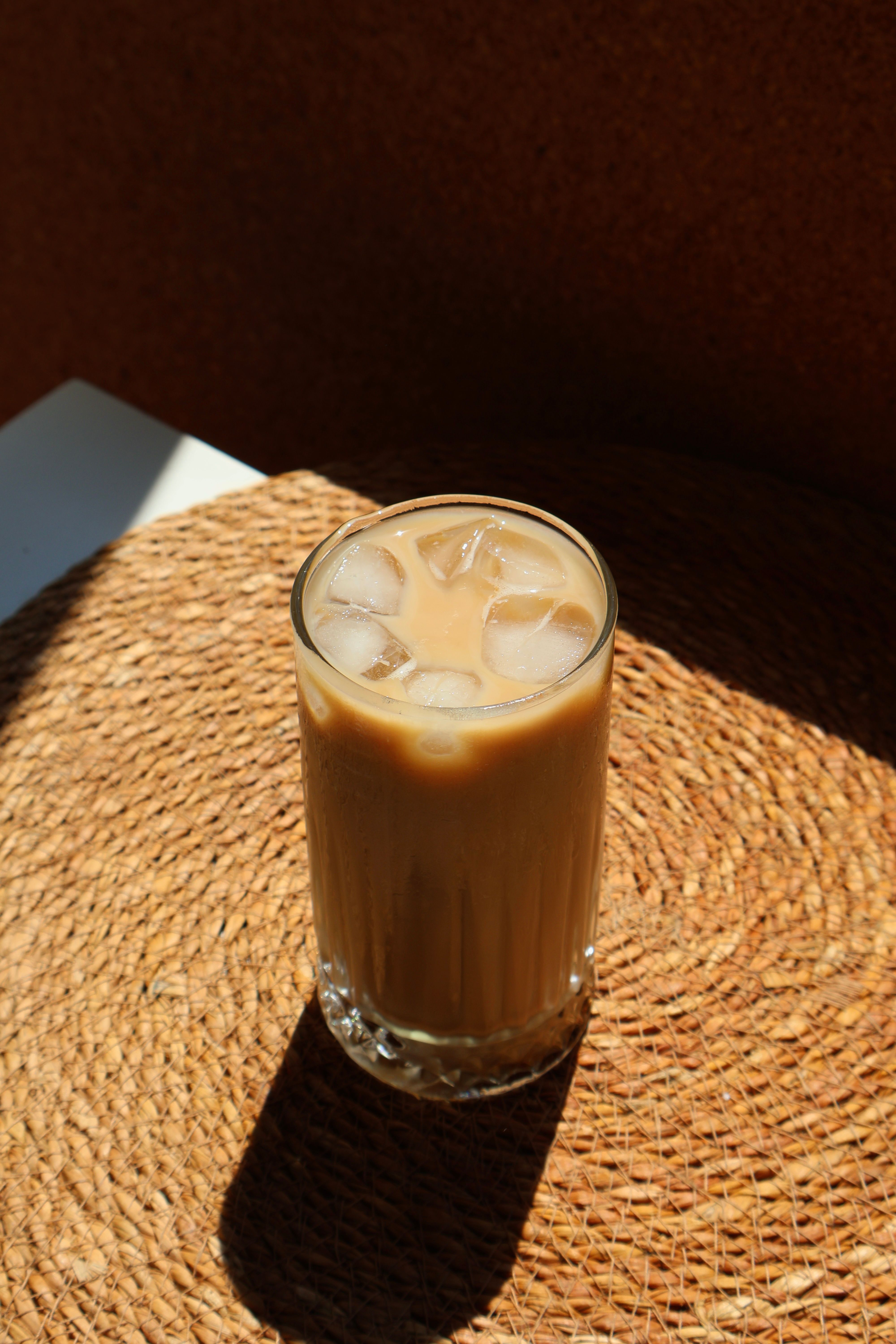 A tall glass of iced coffee with milk sits on a woven mat, bathed in sunlight.