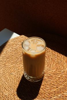 A tall glass of iced coffee with milk sits on a woven mat, bathed in sunlight.