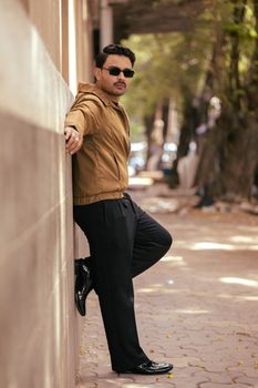 Man wearing sunglasses and casual jacket leaning against a wall in a city street.