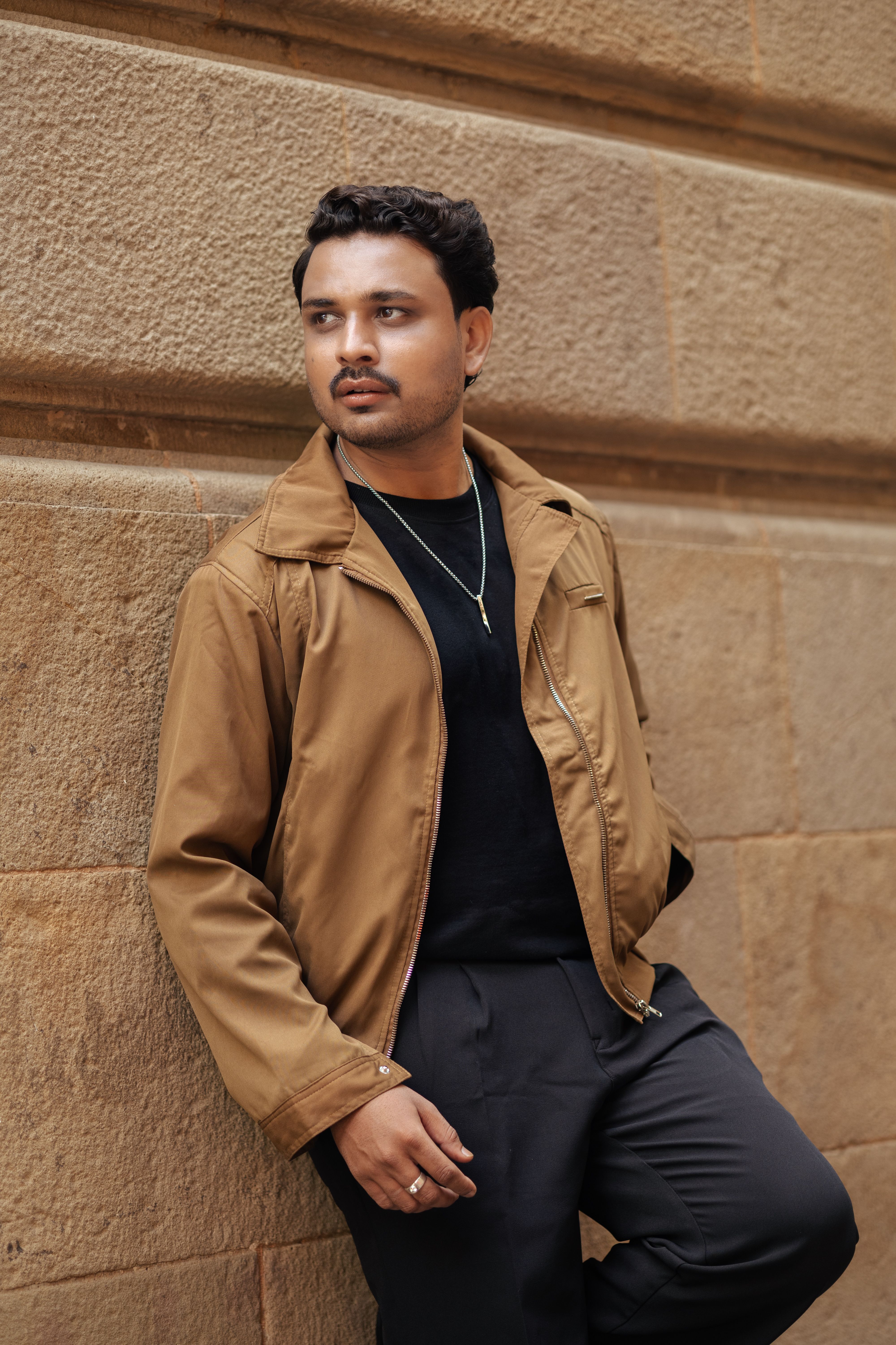 Casual urban fashion with a man leaning on a brick wall wearing a jacket.