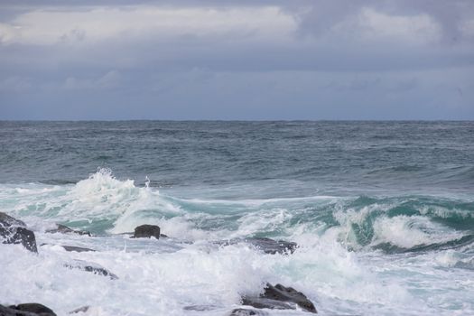 Dynamic ocean waves crashing against rocky shoreline under cloudy sky.