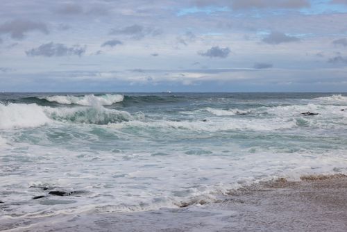 Free Stunning view of ocean waves crashing onto the sandy shore under a cloudy sky. Stock Photo