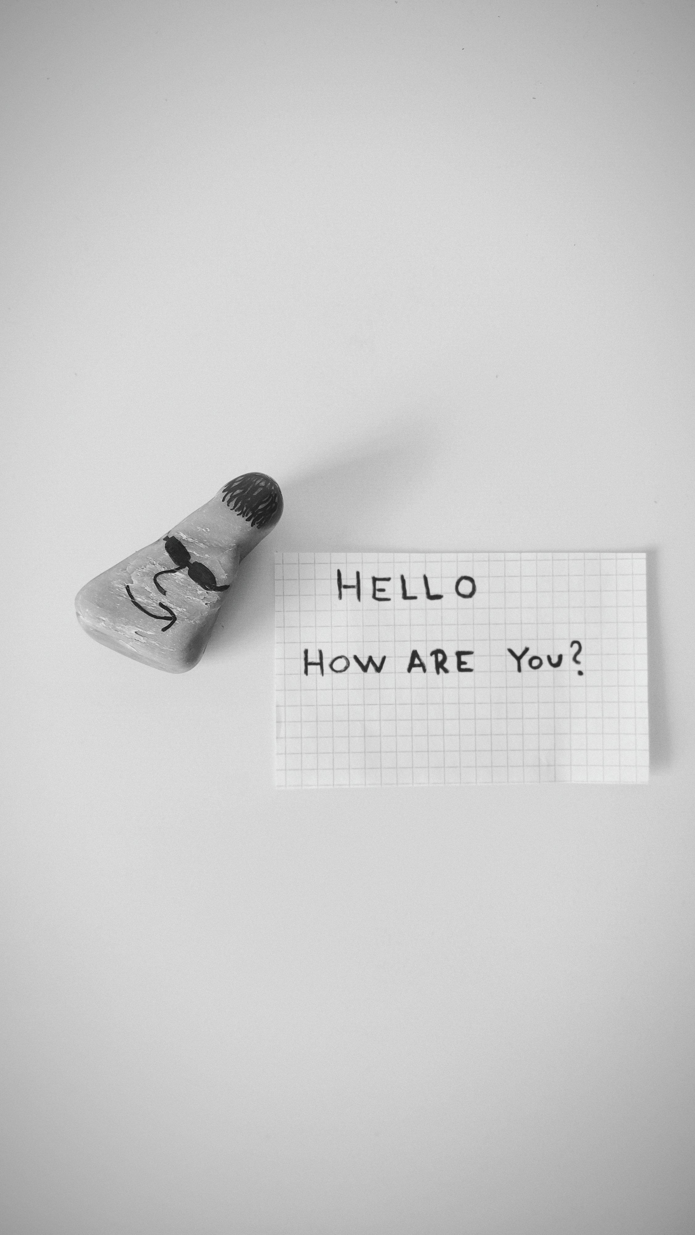Monochrome image featuring a note saying 'Hello, How are you?' beside a humorously painted stone.