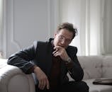 一位穿着黑色西装的自信商人坐在室内沙发上，表情深思熟虑