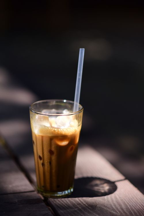 Free A glass of iced coffee with a straw on a wooden table, bathed in sunlight and shadow. Stock Photo