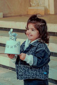 Charming child joyfully holds a birthday cake, featuring a delightful mouse figure and decorations, in an outdoor setting.