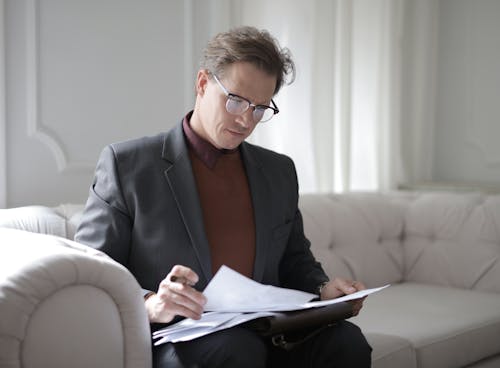 Free Elegant adult man in jacket and glasses looking through documents while sitting on white sofa in luxury room Stock Photo