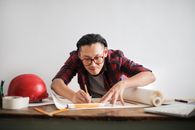 一位受启发的年轻男工程师戴着眼镜，正靠着白墙在桌上绘制草稿