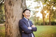 在公园里，一位微笑的亚洲男子穿着西装靠在一棵树上，享受着明媚的一天
