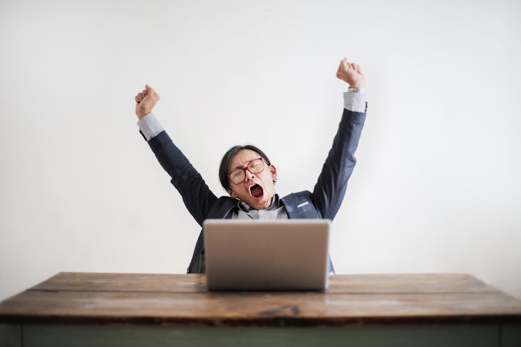 Photo Of Yawning Man With His Hands Up And Eyes Closed Sitting At A Table With His Laptop