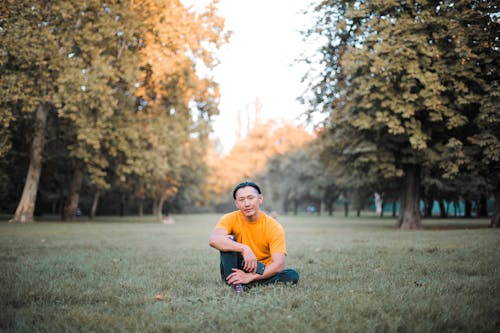 Photo of Man in Orange T-shirt Sitting Alone on Green Grass Field While Holding His Smartphone