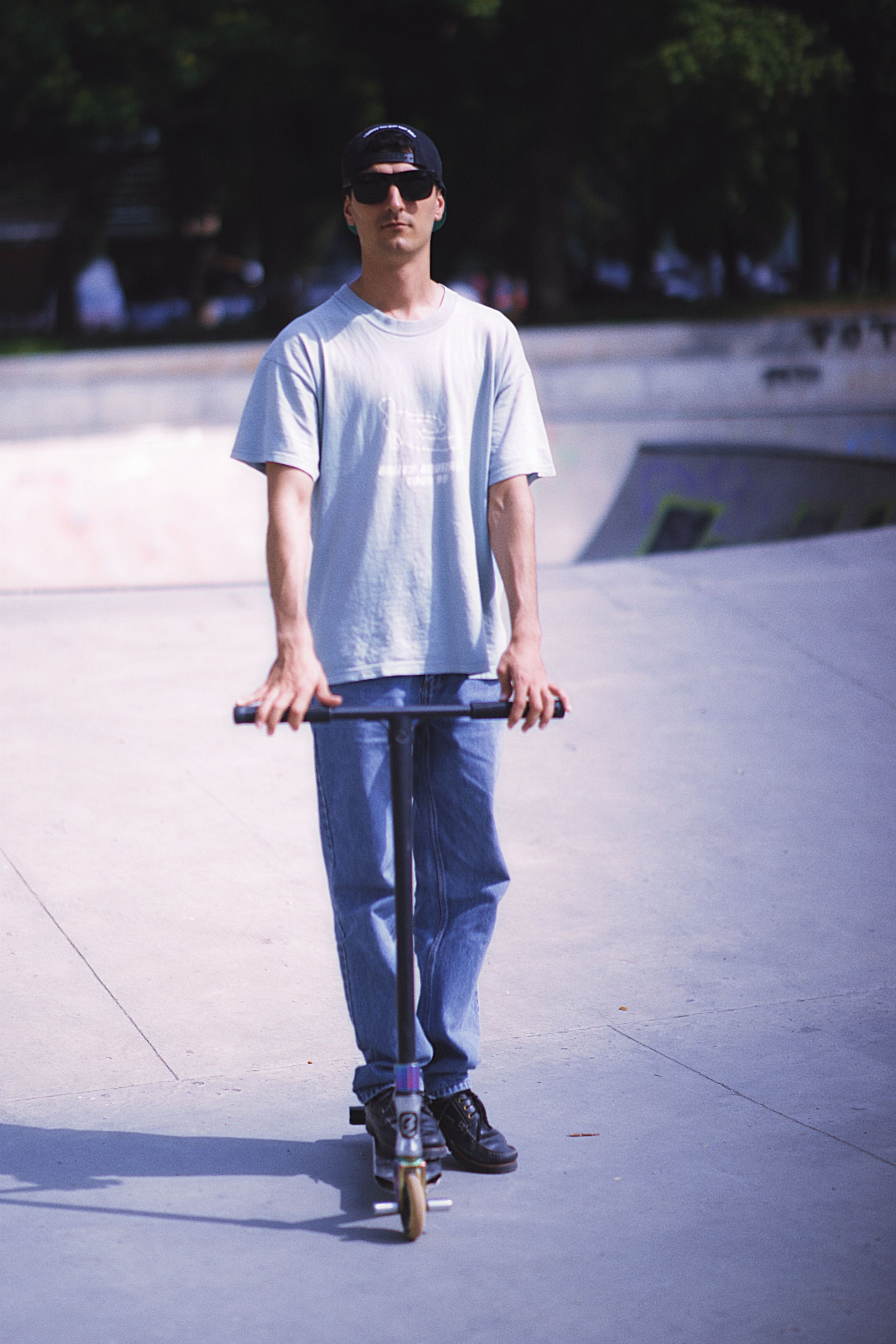 A young man wearing casual clothes rides a scooter in a concrete skate park during the day.
