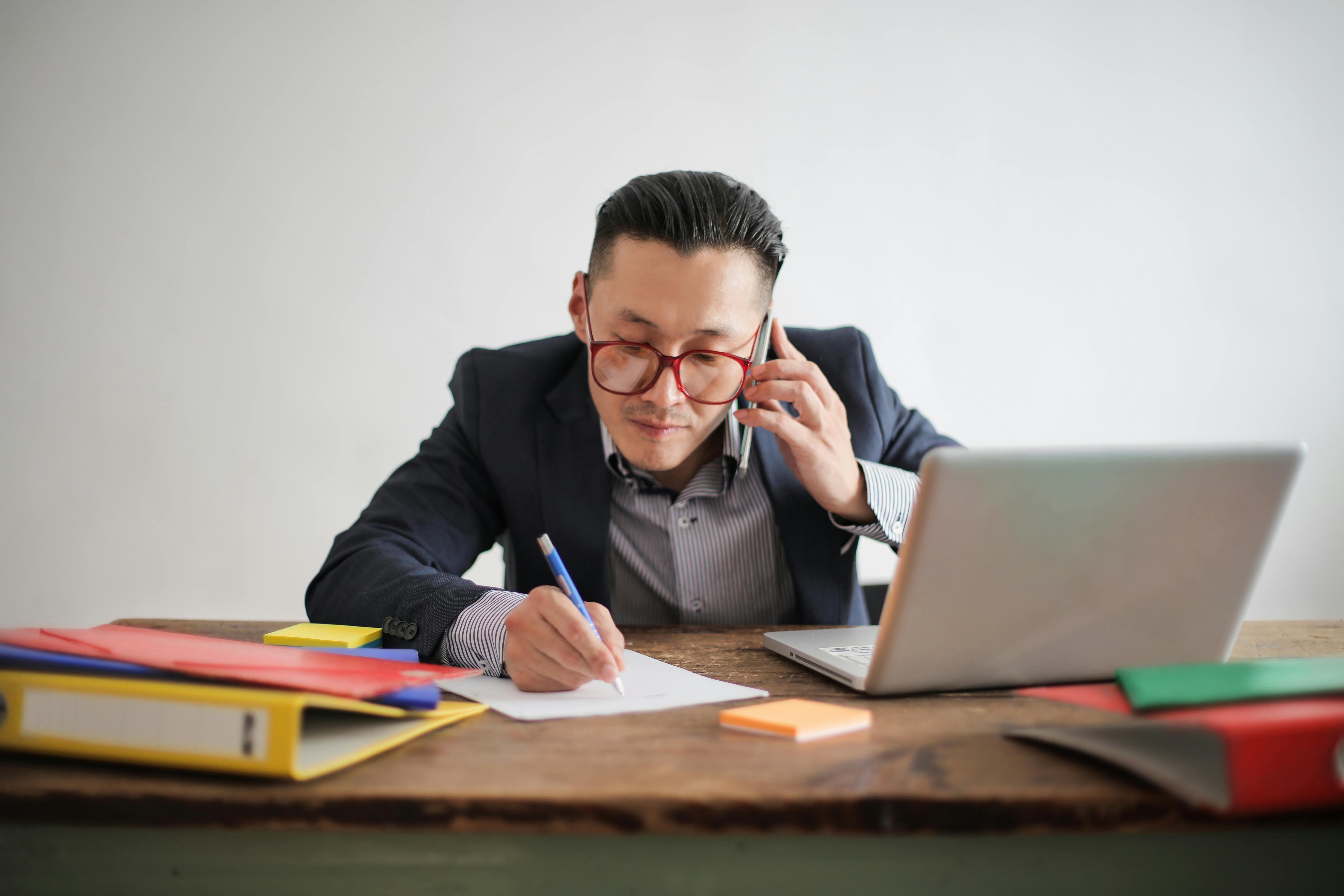 Man talking on the phone while sitting at a table with his laptop. | Photo: Pexels 