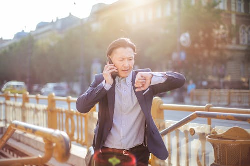 Photo of Man in Blue Blazer and Striped Shirt Standing by Stairs While on the Phone and Looking at His Watch