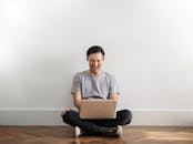 年轻人坐在木地板上，对着白色的墙壁，微笑着用笔记本电脑远程工作