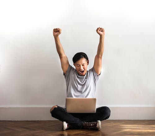 Photo of Man in Gray T-shirt and Black Pants Sitting on Wooden Floor and Working on His Laptop Celebrating with His Hands Raised