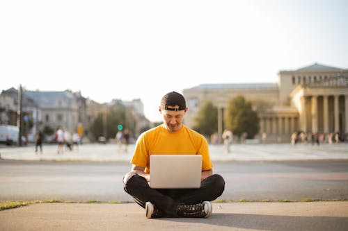 Foto De Foco Seletivo De Homem De Camiseta Amarela E Calça Jeans Preta Sentado No Chão Enquanto Usa Seu Laptop