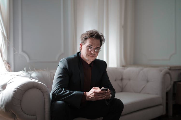 Photo Of Smiling Man In Black Suit Sitting On White Couch Using His Phone