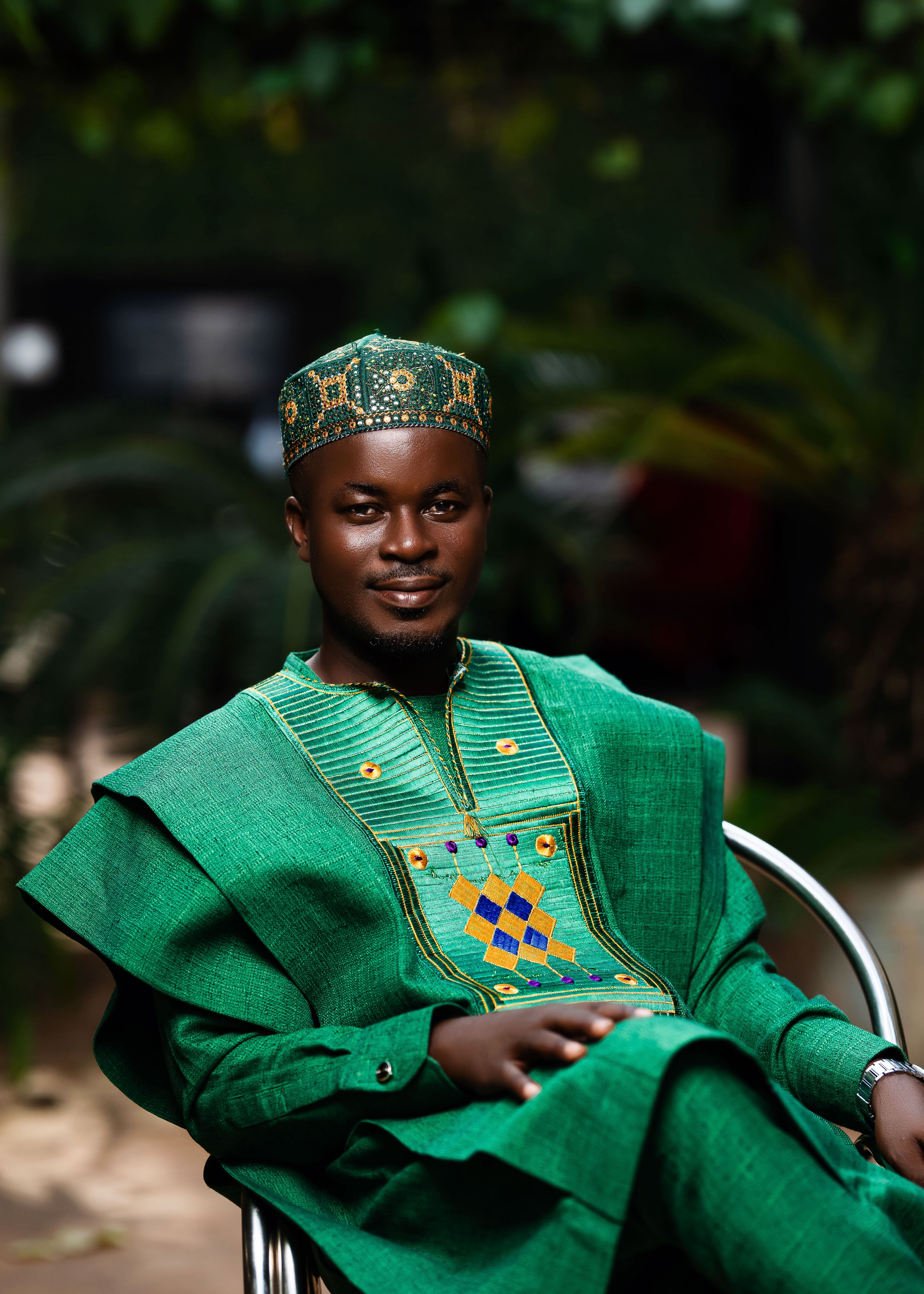 Portrait of a smiling African man in vibrant green traditional attire, seated outdoors with lush greenery.