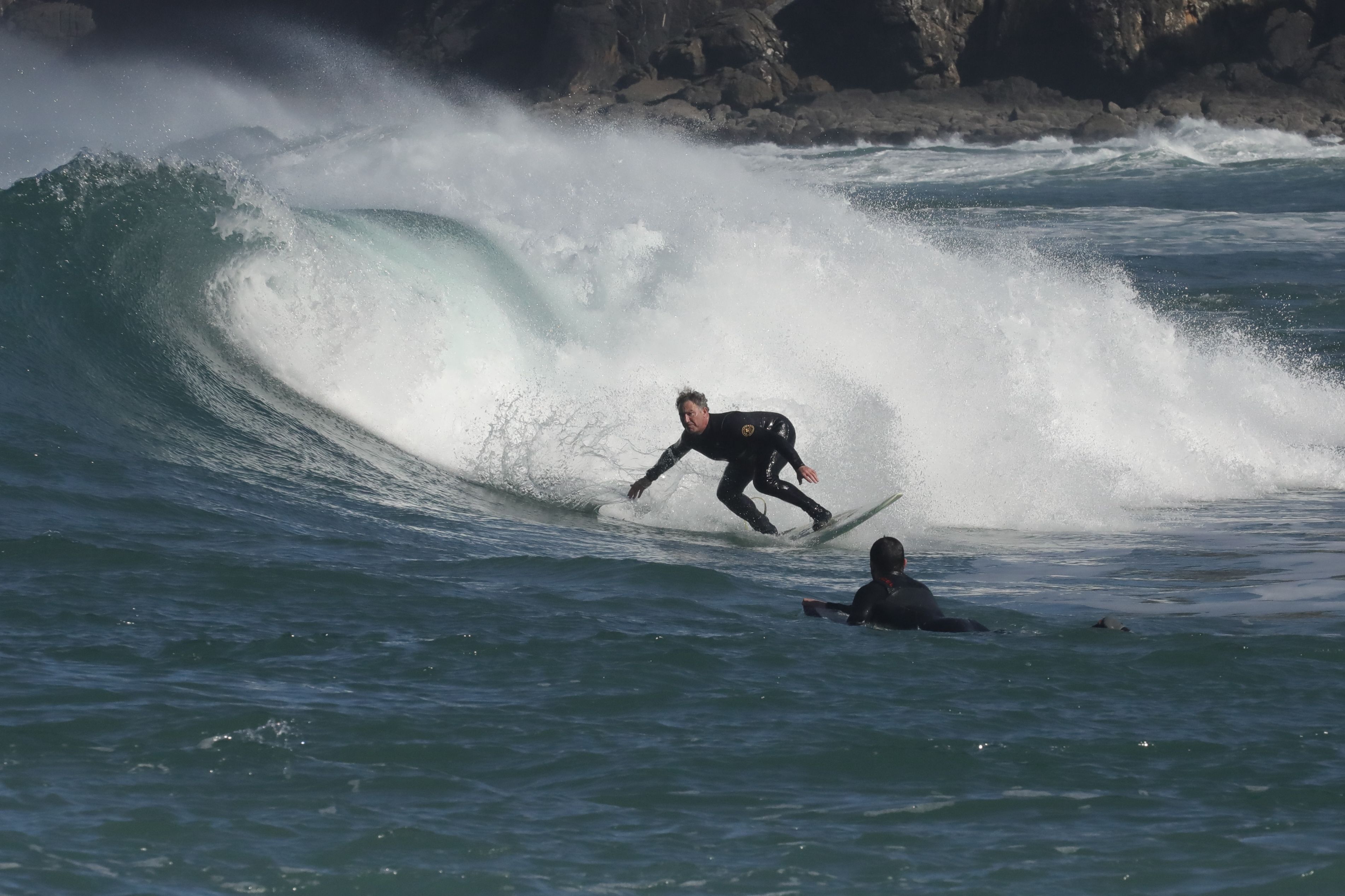 Surfers riding a strong wave near a rocky shoreline, capturing the thrill of the ocean.