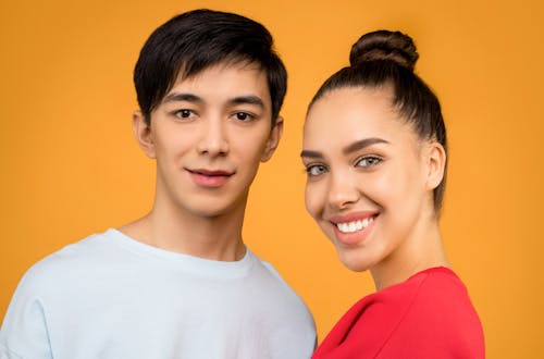 Foto De Retrato De Um Homem Em Uma Camiseta Branca Com Gola Redonda Em Pé Ao Lado De Uma Mulher Sorridente Com Uma Camiseta Vermelha Com Gola Redonda Na Frente De Um Fundo Laranja