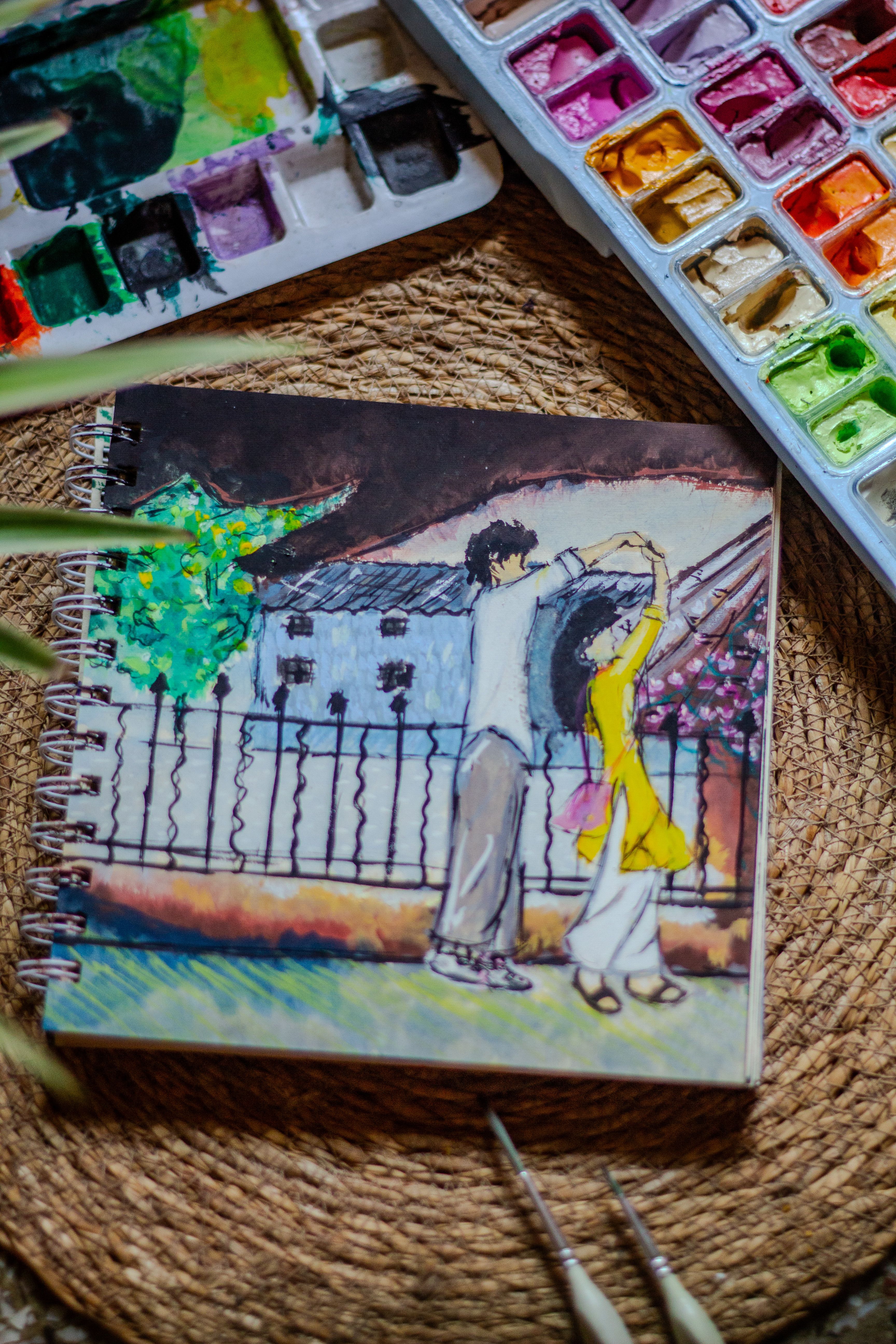 Colorful watercolor notebook with a romantic couple illustration, surrounded by vibrant painting supplies.