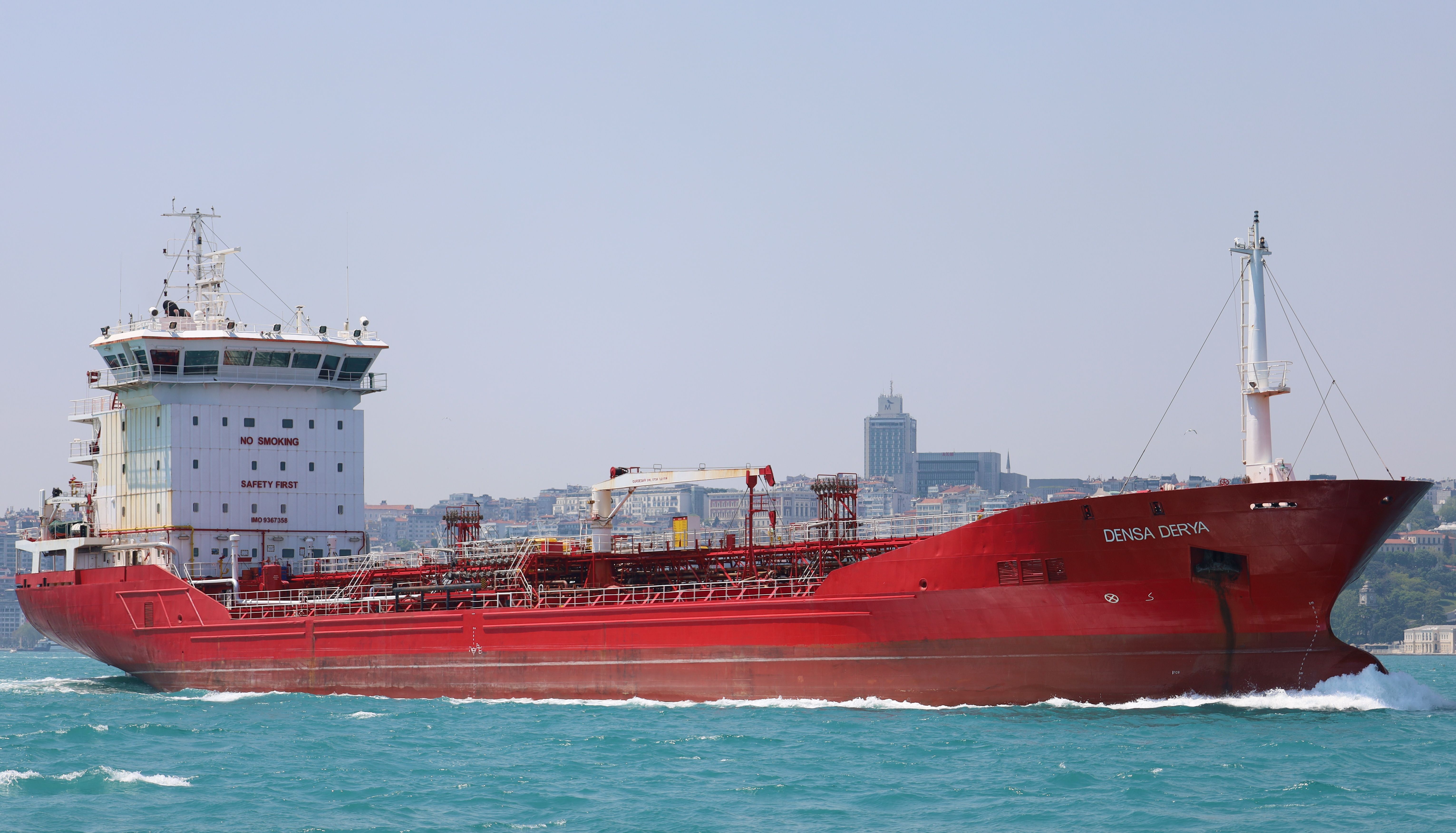 A striking red cargo ship navigating through the blue waters near a cityscape.