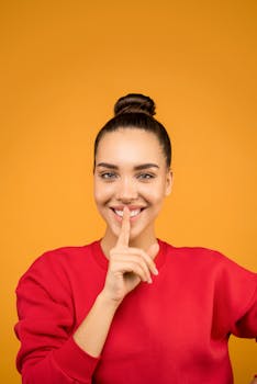 一位年轻女子在生动的黄色背景前，微笑着用手指轻触嘴唇
