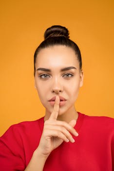 一位优雅的年轻女子在充满活力的黄色背景下，用手指轻触嘴唇摆出姿势