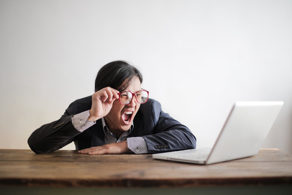 Modern Asian man in jacket and glasses looking at laptop and screaming with mouth wide opened on white background