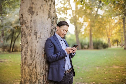 在一个阳光明媚的公园里，一位时尚的年轻人穿着蓝色西装，靠在树上使用智能手机