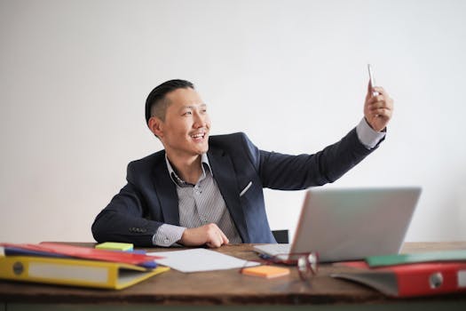 面带微笑的商人在放着笔记本电脑和文件夹的办公桌前用智能手机拍照