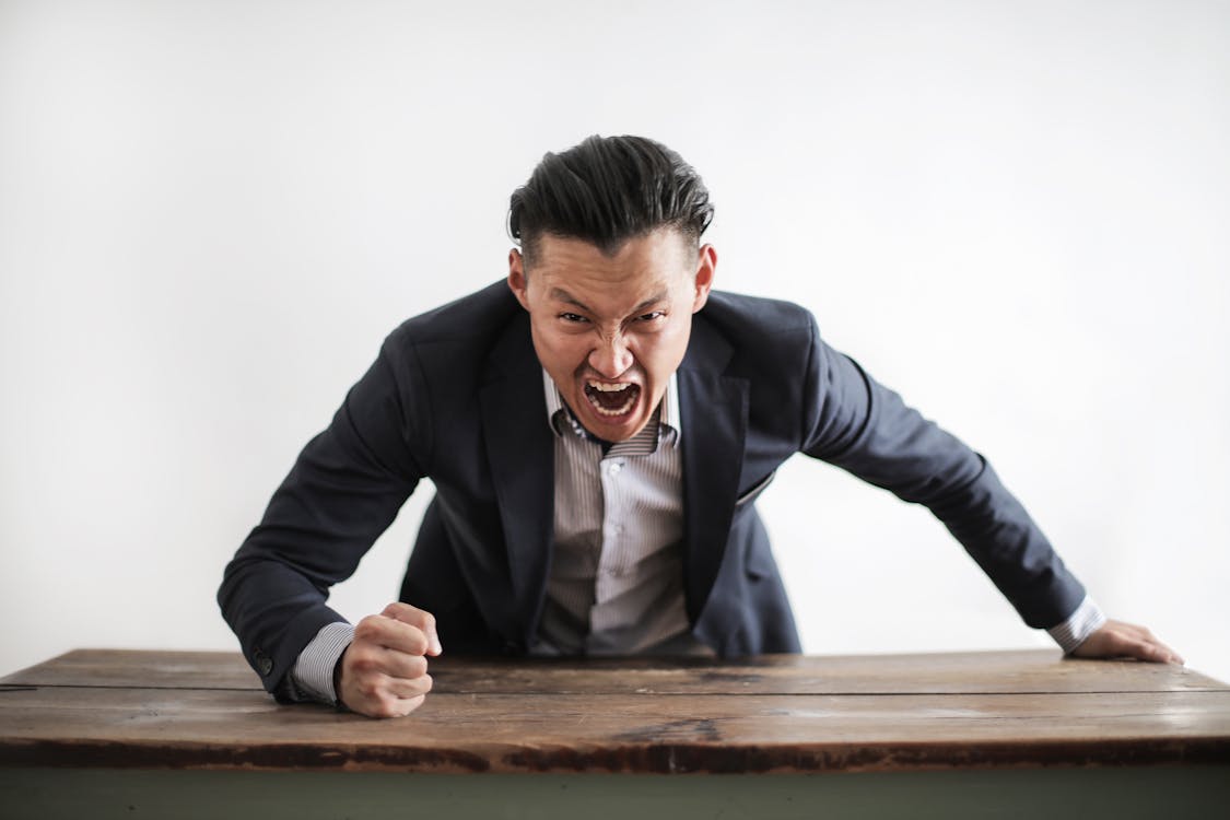 Free Expressive angry businessman in formal suit looking at camera and screaming with madness while hitting desk with fist Stock Photo