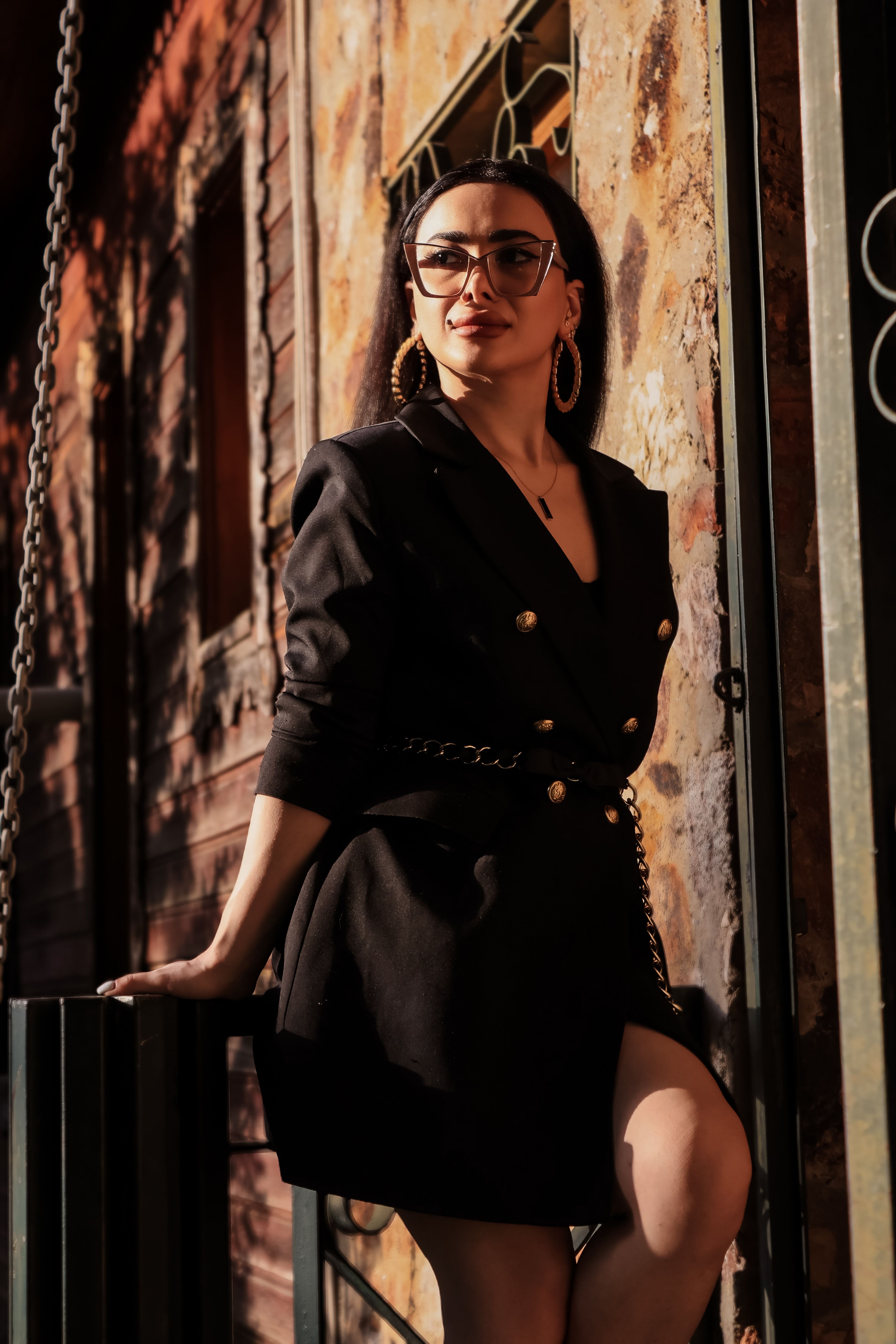 Chic woman in black dress with glasses posing against rustic wall, bathed in warm light.