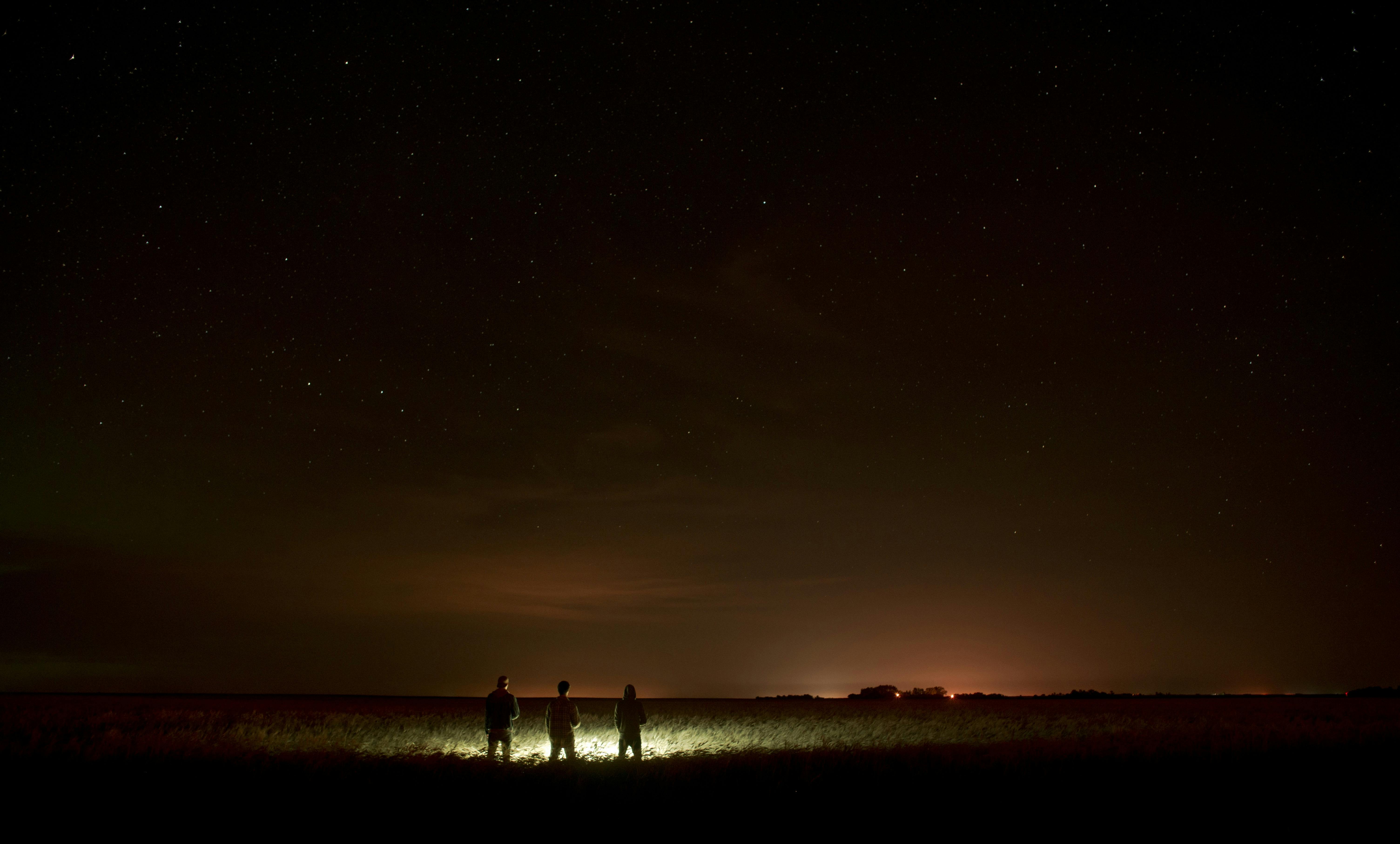 Three people under a starlit sky with distant city lights on the horizon.