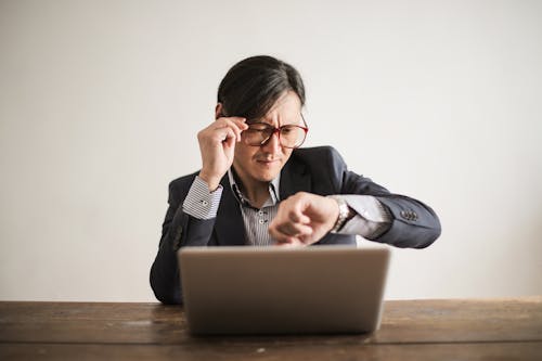 Free Young frowning man in suit and glasses looking at wristwatch while waiting for appointment sitting at desk with laptop Stock Photo