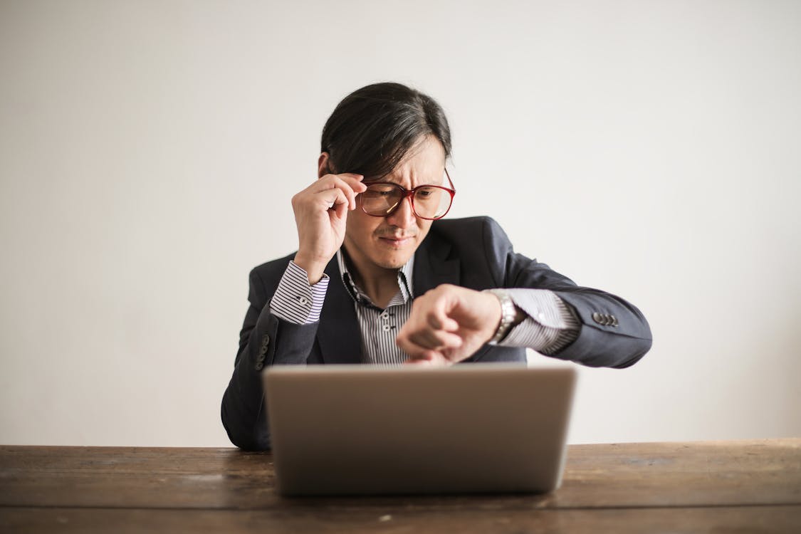 Free Young frowning man in suit and glasses looking at wristwatch while waiting for appointment sitting at desk with laptop Stock Photo