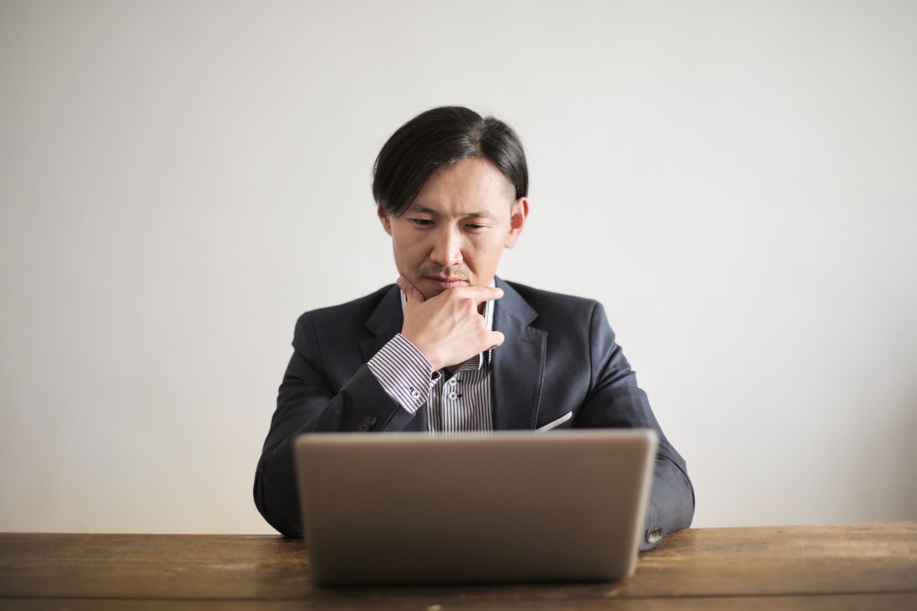 Asian man in suit focusing intently while working on a laptop at a wooden desk indoors.