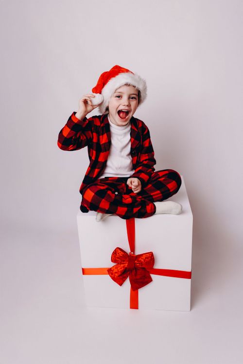 Free A joyful child wearing a Santa hat sits on a large gift box, spreading holiday cheer. Stock Photo