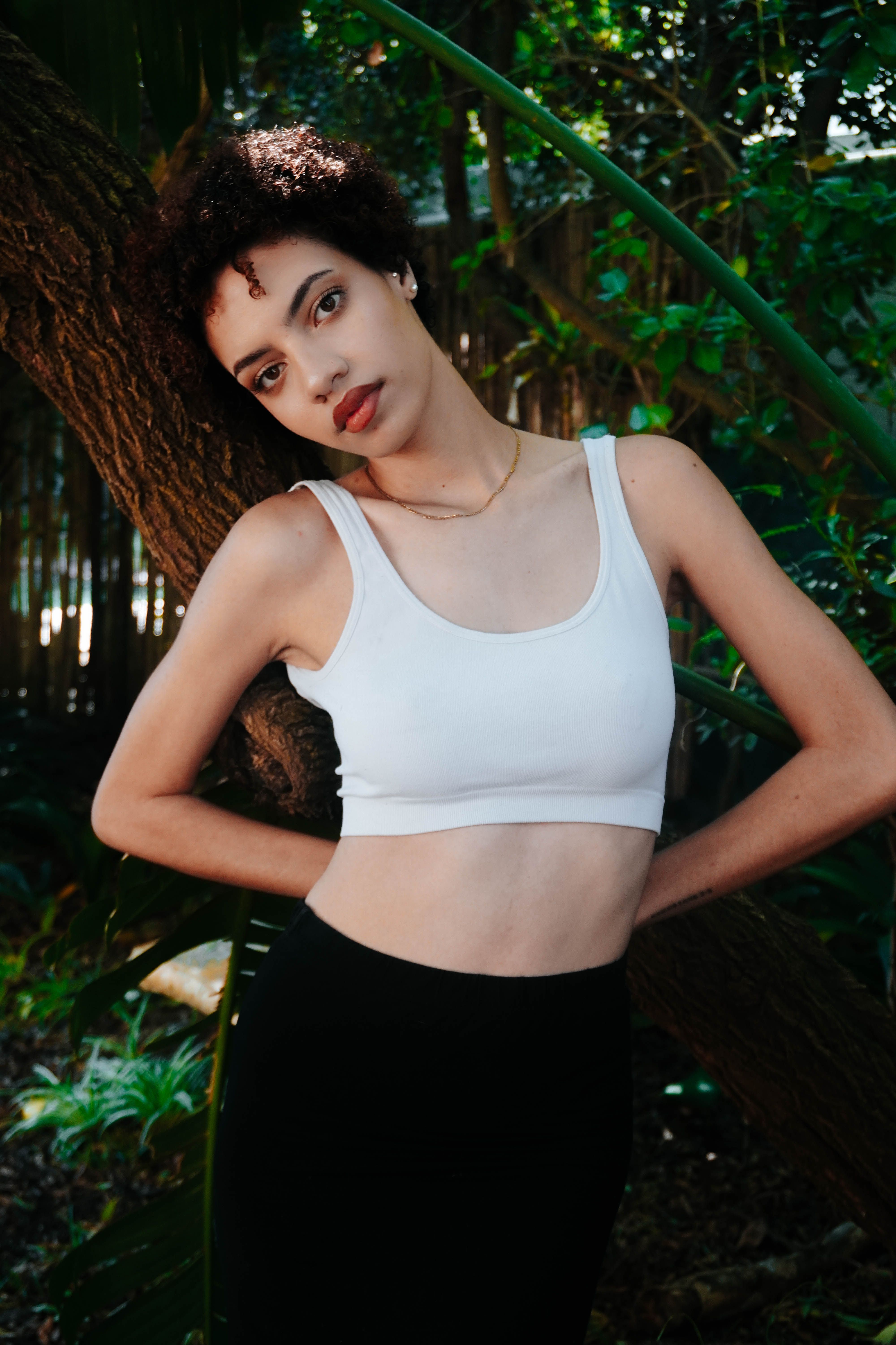Young woman in a white top and black skirt poses outdoors against a tree, surrounded by lush greenery.