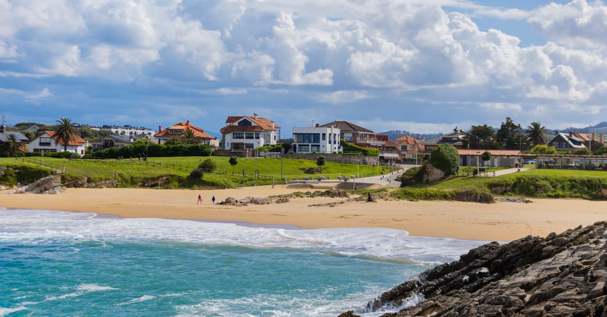 Charming coastal town with residential houses overlooking a sandy beach and rocky shoreline under a blue sky.
