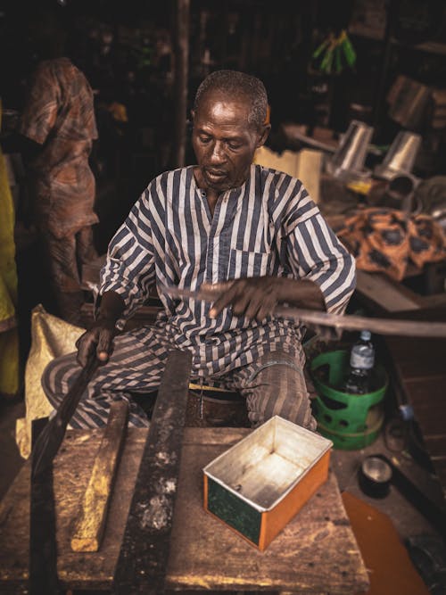 From above of serious middle aged African male artisan in dirty uniform working with metal instruments in old rustic workshop