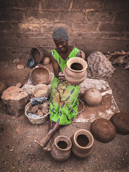 From above of positive barefooted adult African female in authentic clothes and turban sitting on ground and preparing clay pots