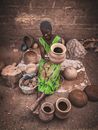 From above of positive barefooted adult African female in authentic clothes and turban sitting on ground and preparing clay pots