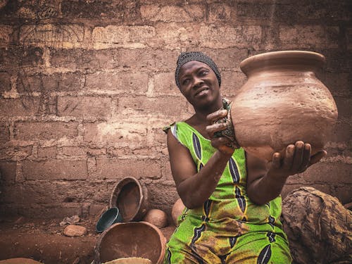 Smiling middle aged ethnic female in traditional dress and turban demonstrating handmade clay pot while sitting on street against shabby wall