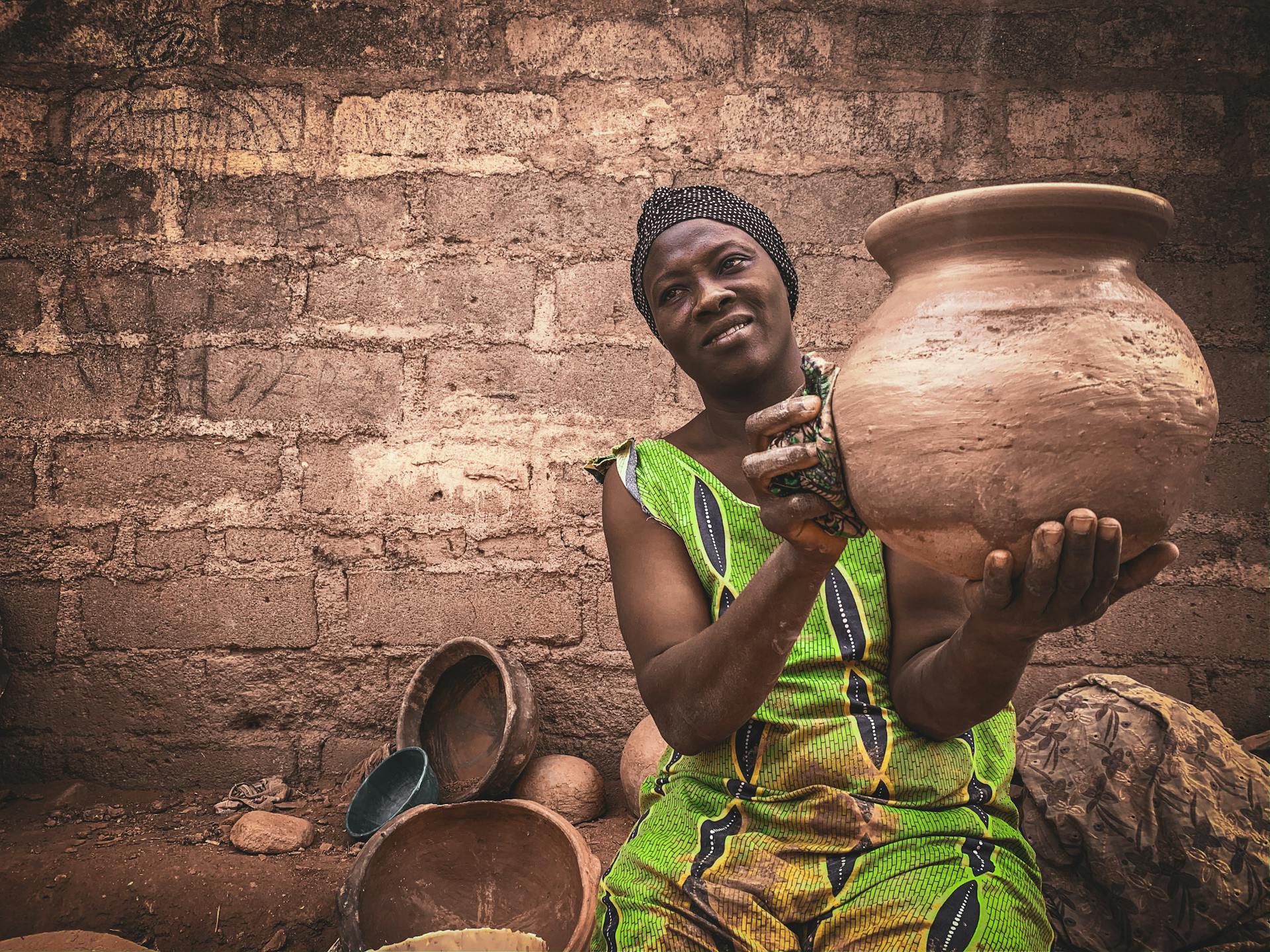 Smiling middle aged ethnic female in traditional dress and turban demonstrating handmade clay pot while sitting on street against shabby wall