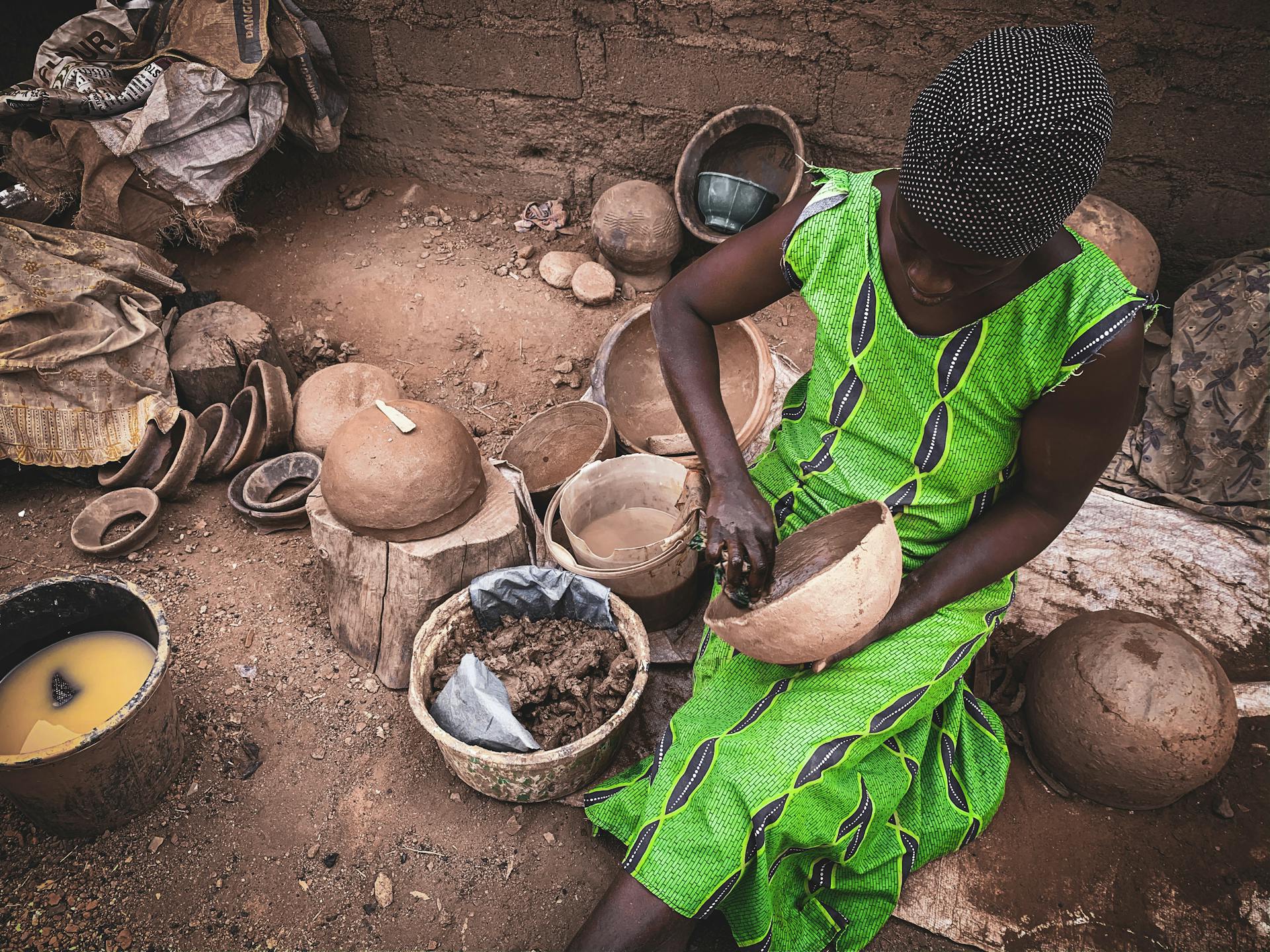 High angle of concentrated African female in light dress sitting on dirty ground of local workshop and creating clay utensils