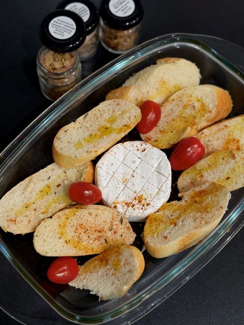 A small glass jar filled with an orange-red all-purpose seasoning blend, a wooden spoon resting on top, with loose paprika and garlic powder scattered on the counter beside it.