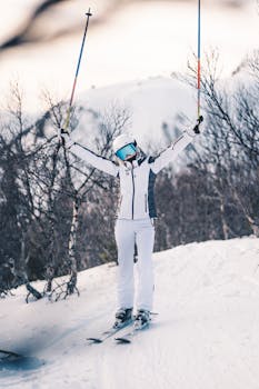 一名身着冬季装备的滑雪者，在被裸露树木包围的白雪覆盖的斜坡上，胜利地举起了滑雪杆