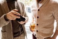 两个穿着休闲毛衣的男人一边展示相机和饮料，一边讨论摄影